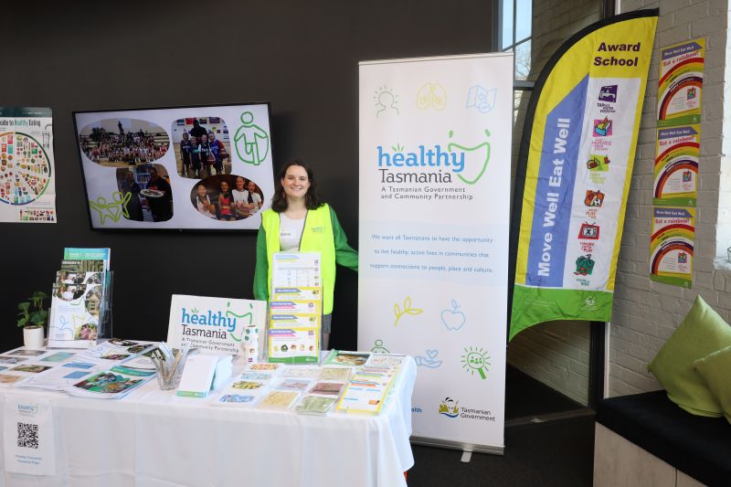 Picture of young girl wearing standing next to the Healthy Tasmania Banner. She is standing at the Healthy Tasmania stall that has a range of pamphlets and resources for guests at the Health Literacy Forum. 