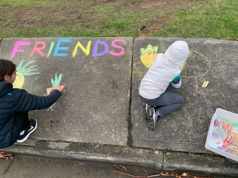 Two children write the word friends in chalk and draw fruit people in chalk messages on a foot path at Mt Nelson.